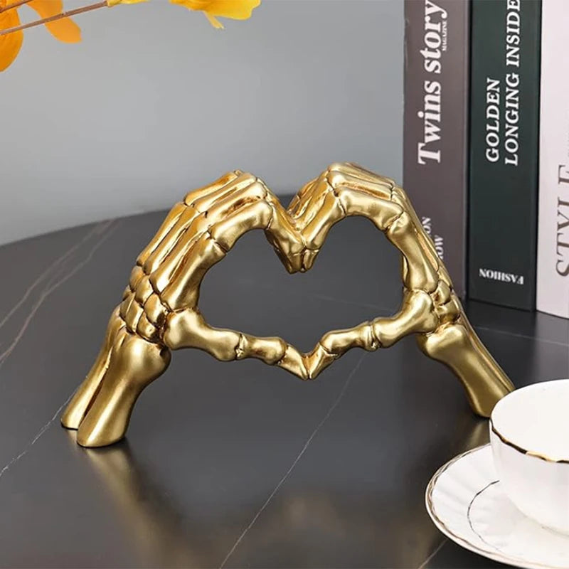 Decorative sculpture of skeletal hands forming a heart shape in gold on a dark surface.